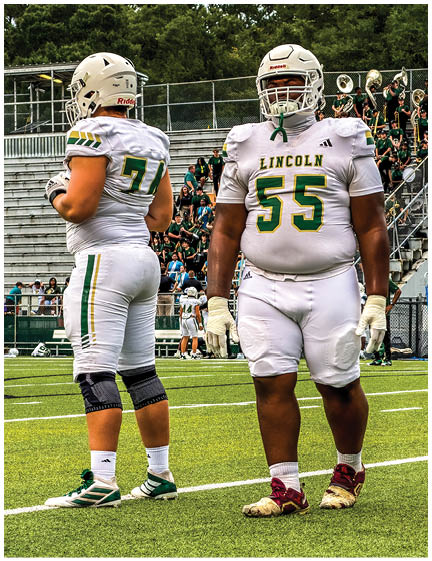 Two people in white football uniforms with green and yellow accents stand on a football field with bleachers and spectators in the background. Description generated by AI