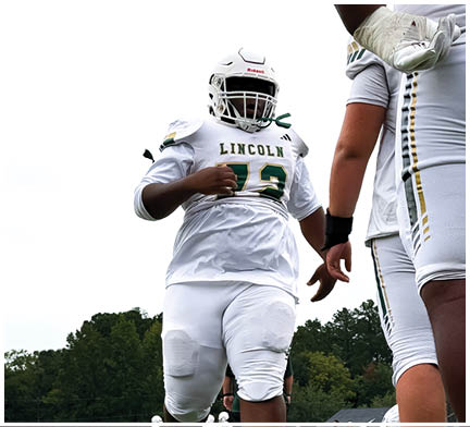 Three people wearing white football uniforms with helmets are standing, with one person in the foreground and two others partially visible. Description generated by AI