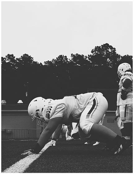 A person wearing a helmet and sports uniform is bent over on a field with trees in the background. Description generated by AI