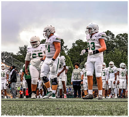 Three people wearing white football uniforms and helmets stand on a field with trees and a cloudy sky in the background. Description generated by AI