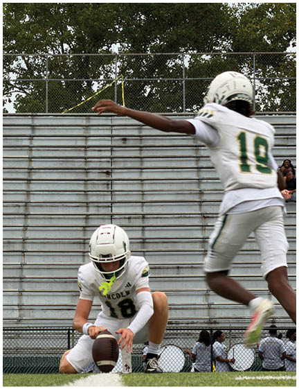 Two people in white football uniforms with the number 18 are on a field, one holding a football in a kneeling position while the other is running nearby with an arm extended. Description generated by AI