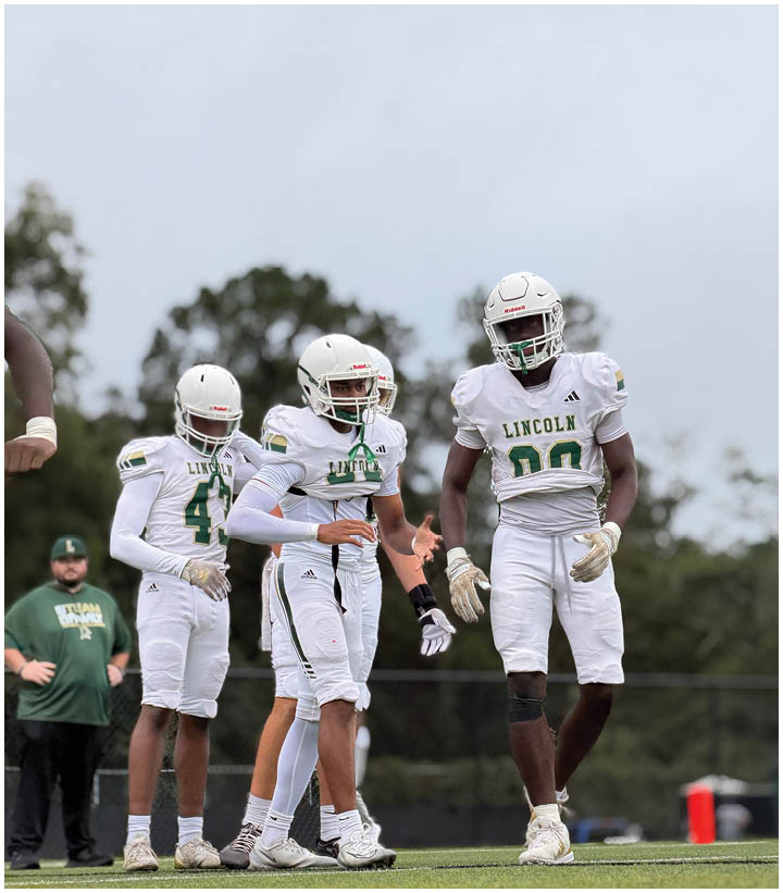 Three people in white football uniforms and helmets stand on a field with a person in a green shirt in the background. Description generated by AI