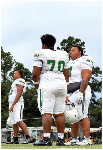 Three people wearing white football uniforms with green and gold accents stand outdoors near trees and a fence, with one holding a helmet. Description generated by AI