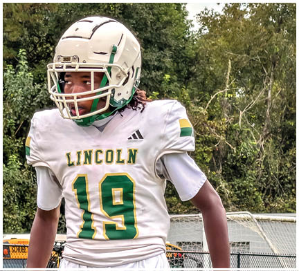 A person wearing a white football helmet and a white jersey with green and yellow accents and the word Lincoln and the number 19 stands outdoors near a football goalpost with trees in the background. Description generated by AI
