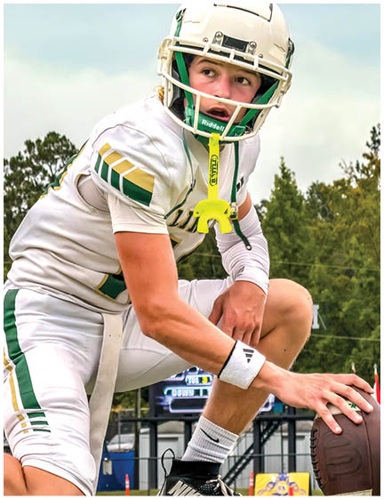 A person wearing a white football uniform and helmet is crouched down holding a football on a field with trees and a scoreboard in the background. Description generated by AI
