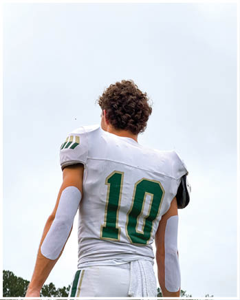 An adult wearing a dark head covering and shirt is crouched in a ready position on a sports field with another person in green sports gear blurred in the background. Description generated by AI