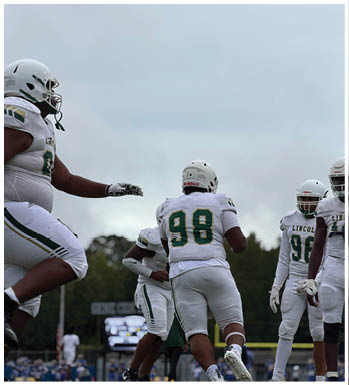 An adult wearing a dark head covering and shirt is crouched in a ready position on a sports field with another person in green sports gear blurred in the background. Description generated by AI