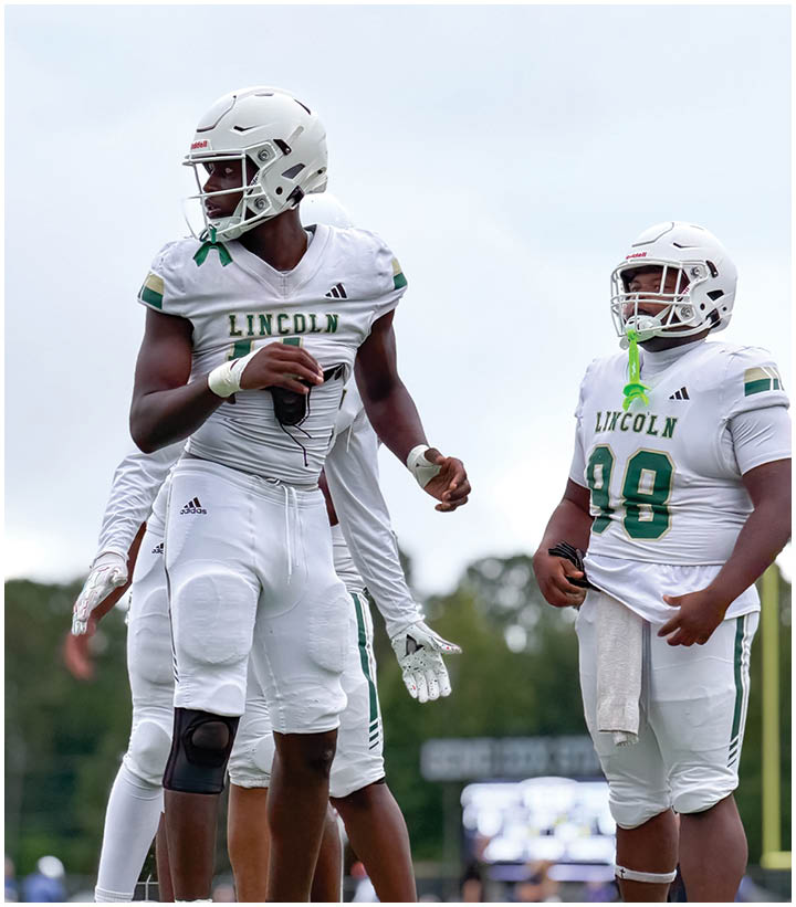 Three people wearing white football uniforms with “LINCOLN" on the jerseys and helmets stand on a field, with one person holding a football and another with a mouthguard hanging from the helmet. Description generated by AI