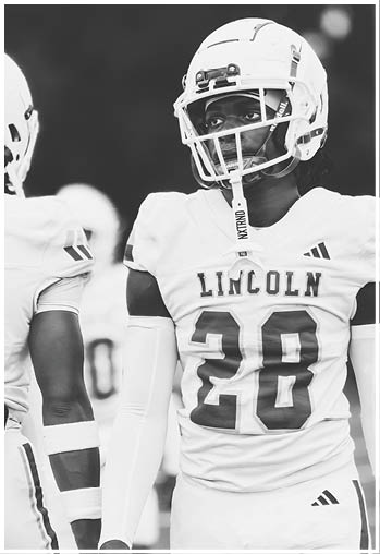 Two people wearing football helmets and uniforms with the word Lincoln and the number 28 on one jersey are standing on a field. Description generated by AI