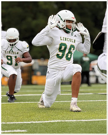 Two people in white football uniforms with “LINCOLN" on the jerseys are on a field, with one kneeling and raising their hands near their helmet while the other is running behind. Description generated by AI