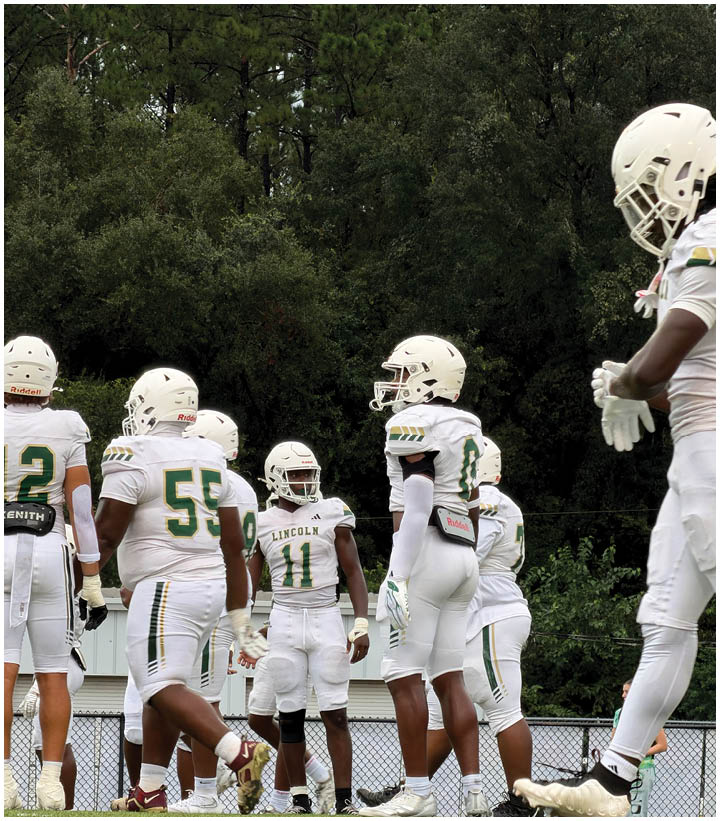 A group of people wearing white football uniforms and helmets are standing on a field with trees in the background. Description generated by AI