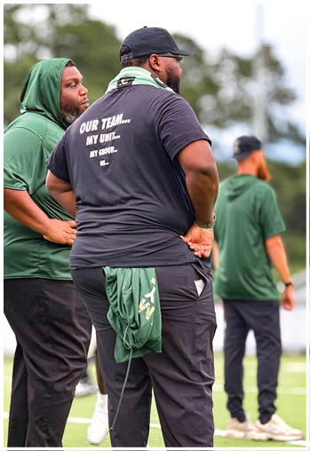 Three adults wearing casual sportswear stand on a field with one person in the foreground wearing a black shirt with text on the back and a green garment tucked into their waistband. Description generated by AI
