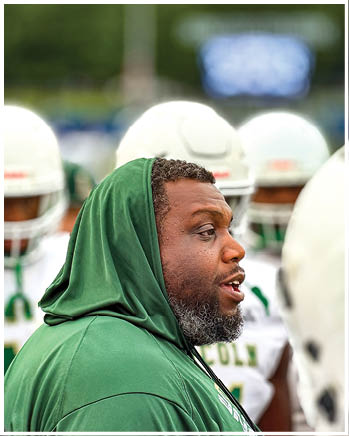 A person wearing a green hooded sweatshirt is speaking on the sidelines with several people in white football helmets and uniforms in the background. Description generated by AI