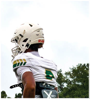 An adult wearing a dark head covering and shirt is crouched in a ready position on a sports field with another person in green sports gear blurred in the background. Description generated by AI