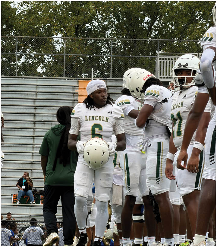 People in football uniforms stand on a field near empty bleachers, with one person holding a helmet and others adjusting their gear. Description generated by AI