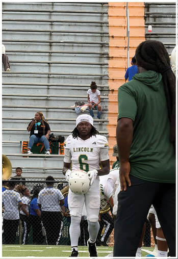A person in a green shirt and black pants stands on a football field facing a group of football players in white uniforms and helmets with one player holding a helmet walking toward them, with empty bleachers and a few seated people in the background. Description generated by AI