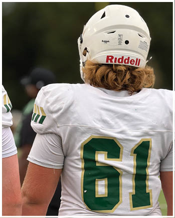 A person wearing a white football jersey with the number 61 and a white helmet stands on a field with blurred people in the background. Description generated by AI