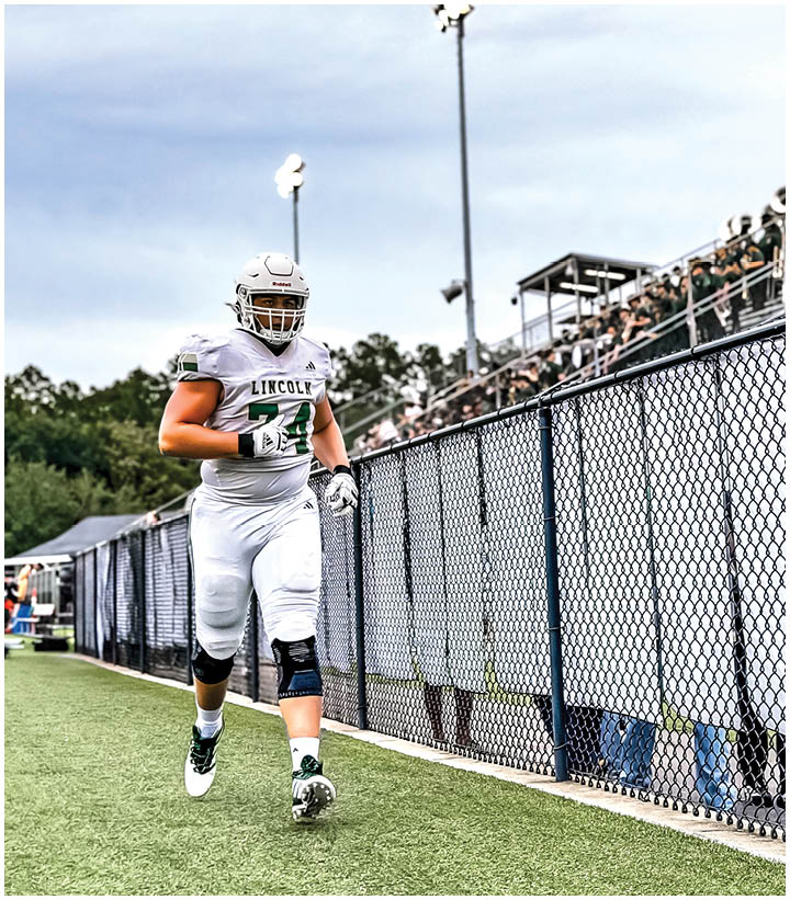 An adult in a white football uniform with a helmet is walking on a field near a chain-link fence with spectators in bleachers in the background. Description generated by AI