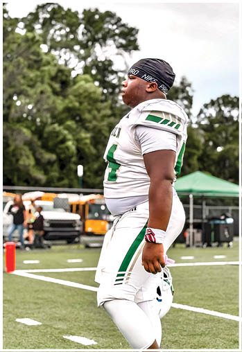 A person wearing a white and green football uniform and a black headband is standing on a football field with trees and vehicles in the background. Description generated by AI