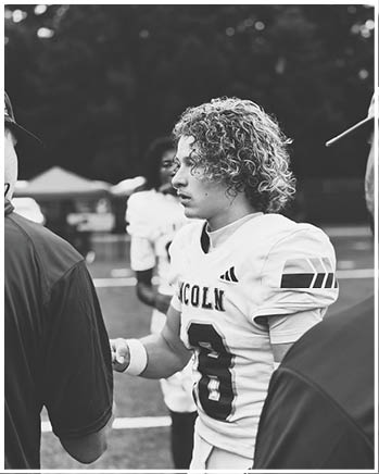 A person with curly hair wearing a football uniform with the number 8 and the word Lincoln is standing on a field talking to two other people. Description generated by AI