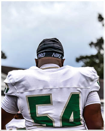 A person wearing a white sports jersey with the number 54 in green and a black Nike head covering is seen from behind outdoors. Description generated by AI