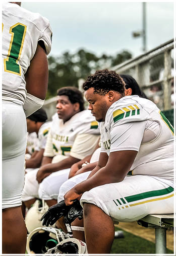 People in football uniforms are sitting on a bench, with one person in the foreground looking down while holding gloves. Description generated by AI