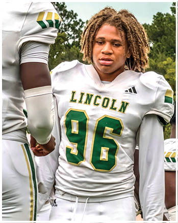 Two people wearing white football uniforms with green and yellow accents are standing outdoors, with one person wearing a jersey labeled “LINCOLN 96" and looking down. Description generated by AI