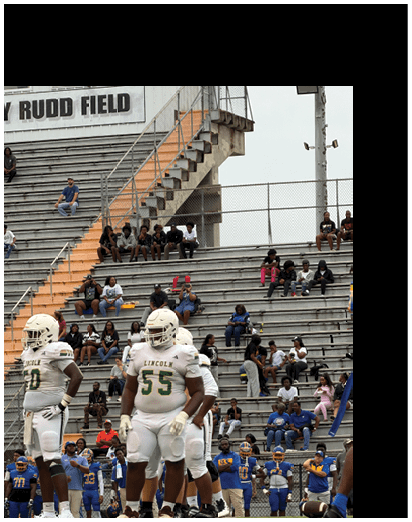 Football players in white uniforms stand on the field while people sit in the bleachers of a stadium. Description generated by AI