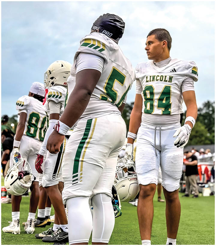 Four people in white football uniforms with green and yellow accents stand on a grassy field, two holding helmets and one wearing a black head covering. Description generated by AI