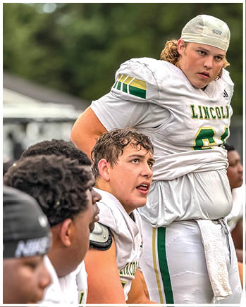 Four people in football uniforms are gathered closely, with one standing and the others sitting, appearing focused and engaged during a game or practice. Description generated by AI