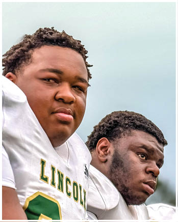 Two people wearing white football uniforms with “LINCOLN" written on the front are sitting closely together, looking thoughtful. Description generated by AI