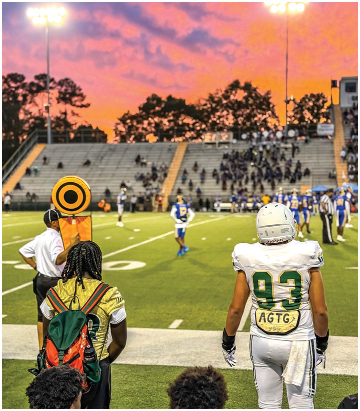A person in a white football uniform with the number 93 stands on the sideline of a football field at sunset while other people and players are visible in the background. Description generated by AI