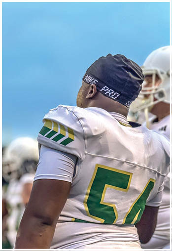 A person wearing a white and green football uniform with the number 54 and a black Nike Pro headband is standing on a field with other players in the background. Description generated by AI