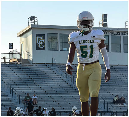 An adult wearing a white football uniform with the number 51 and the word Lincoln walks on a field with empty bleachers and a press box in the background. Description generated by AI