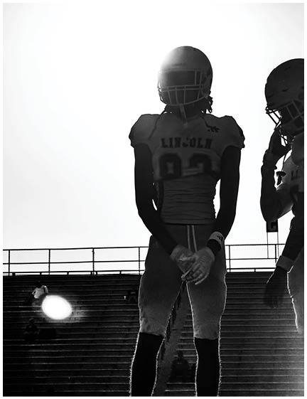 Two people wearing football uniforms and helmets stand on a field with bleachers in the background, one facing forward with hands clasped and the other turned to the side. Description generated by AI