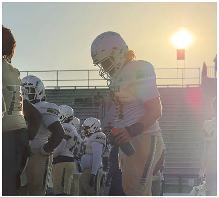 A group of people wearing football uniforms and helmets stand on a field with bleachers in the background, one person in the foreground holding a water bottle. Description generated by AI