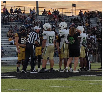 A group of people including football players in uniforms and a referee are gathered on a field near the center with spectators seated in bleachers in the background during sunset. Description generated by AI