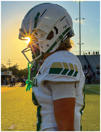 A person wearing a white football uniform and helmet stands on a field at sunset with the sun shining through the helmet's faceguard. Description generated by AI