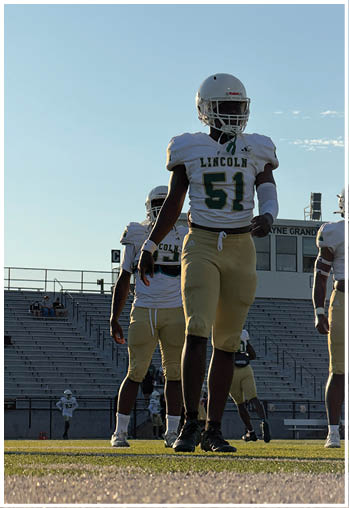 Three people in football uniforms with helmets walk on a field with empty bleachers and a building in the background during daylight. Description generated by AI