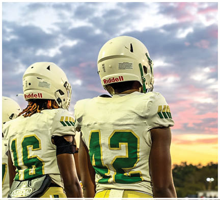 Two people wearing white football uniforms with green and yellow numbers stand side by side under a cloudy sky at sunset. Description generated by AI