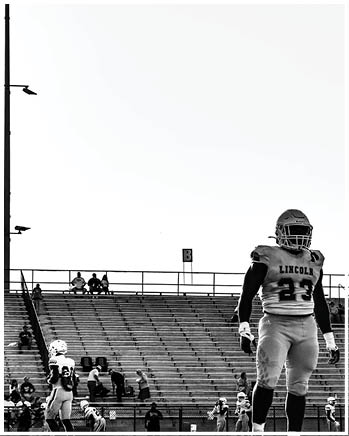 A person in a football uniform with the word Lincoln on the jersey stands on a field with other players and a mostly empty stadium in the background. Description generated by AI
