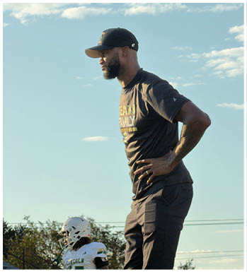 An adult wearing a black cap and black clothing stands with hands on hips watching a child in a white football uniform and helmet on a sports field. Description generated by AI