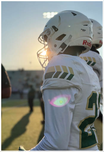 Two people wearing white football helmets and white jerseys with green and gold accents stand on a field with the sun setting behind them. Description generated by AI