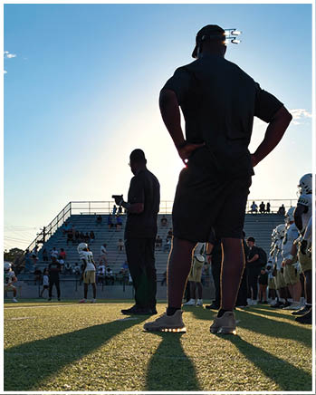 Two adults stand on a football field near players in helmets and uniforms with a stadium and spectators in the background during a clear day. Description generated by AI