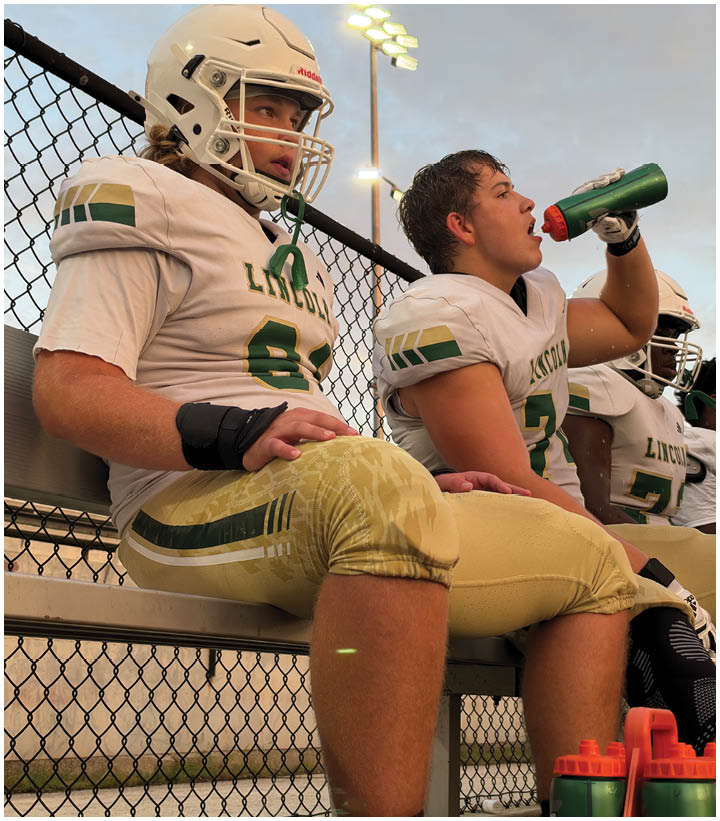 Three people in football uniforms sit on a bench by a chain-link fence, with one person wearing a helmet and another drinking from a green water bottle. Description generated by AI