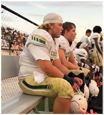 People in football uniforms sit on a bench on the sidelines of a field with helmets and water bottles nearby. Description generated by AI