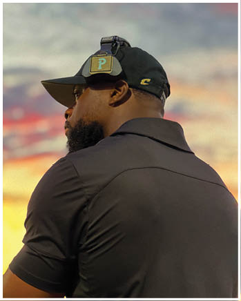 An adult wearing a dark head covering and shirt is crouched in a ready position on a sports field with another person in green sports gear blurred in the background. Description generated by AI