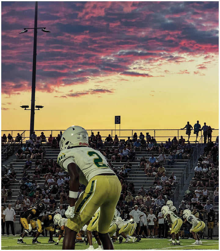 People in football uniforms are lined up on a field preparing for a play with a crowd watching from bleachers under a colorful sunset sky. Description generated by AI