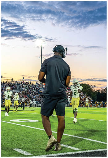 A person wearing a headset and black sportswear stands on the sidelines of a football field while players in yellow and white uniforms are on the field during a sunset game. Description generated by AI