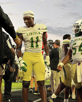 A person wearing a Lincoln football uniform with the number 11 is shaking hands with another person on the sidelines of a football field while other players and people stand nearby. Description generated by AI